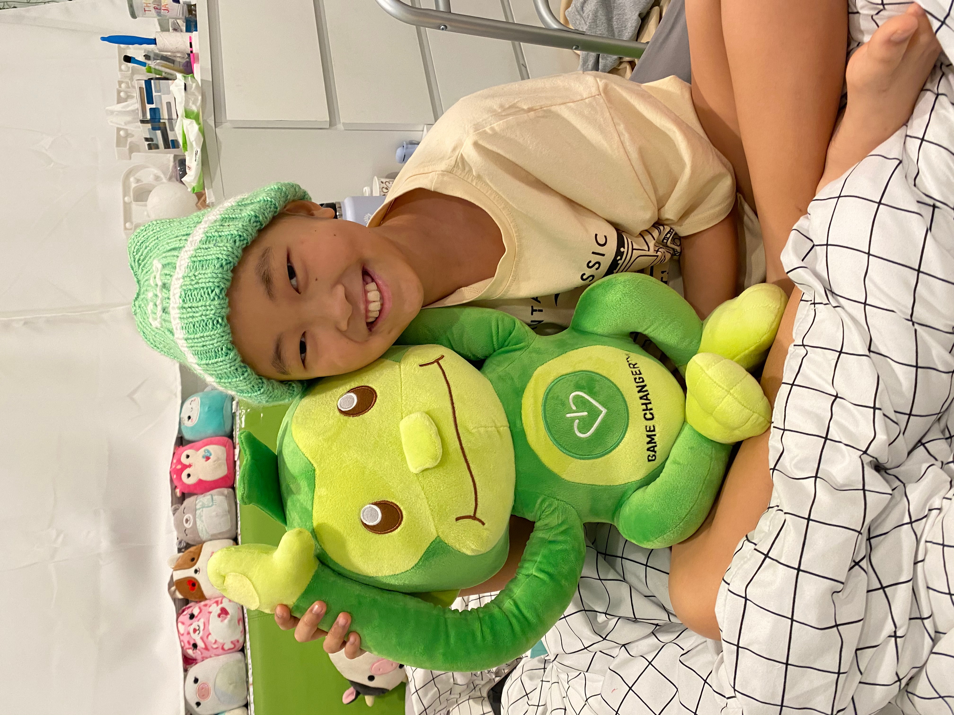 A young patient in a green beanie beaming as she hugs Hope, GameChanger's green monkey.